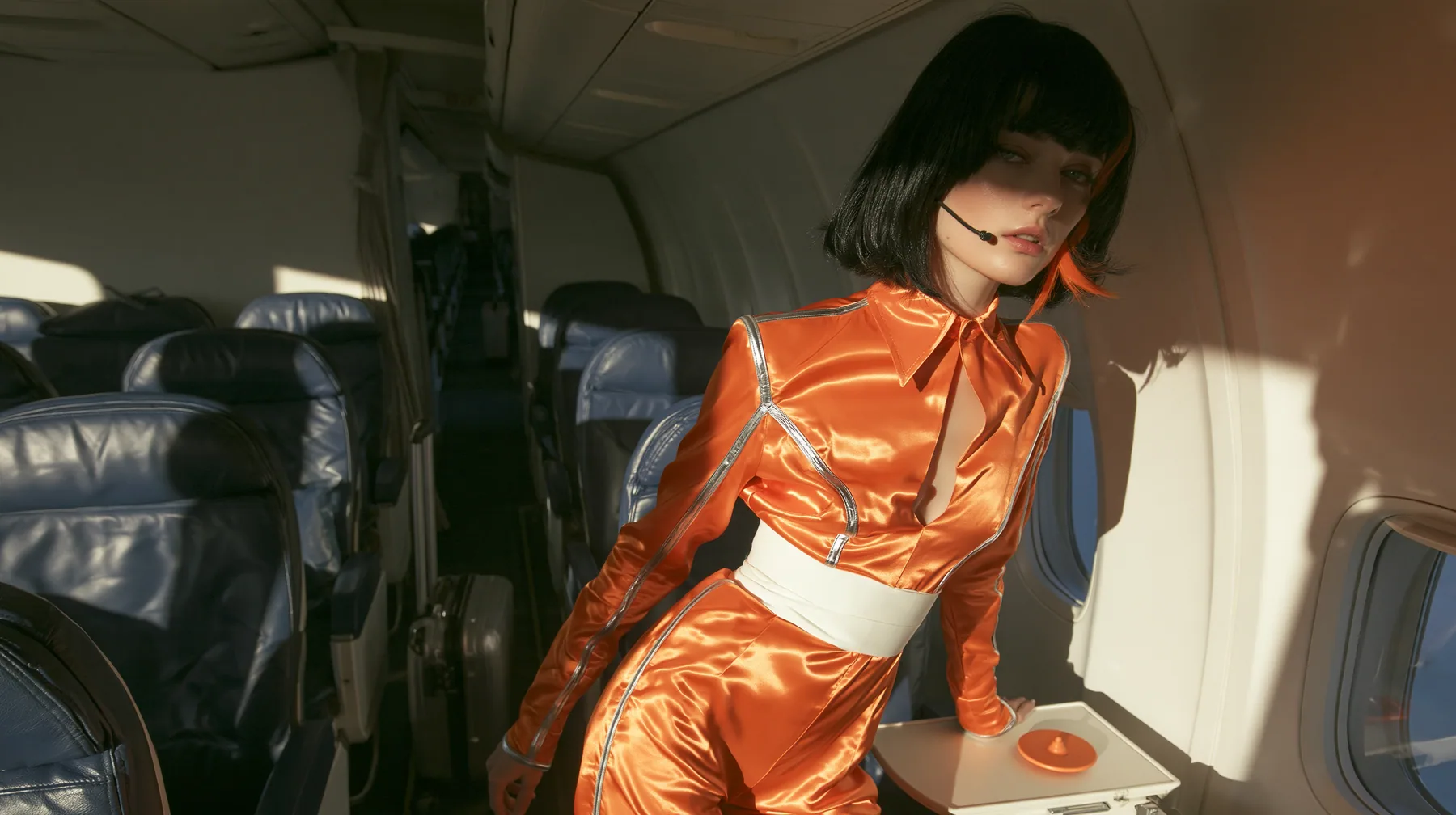 Fashion editorial, sunrise: an eye-level wide shot down the aisle of a widebody airliner cabin with navy leather seats and white walls, golden morning light, a Caucasian American woman in her late twenties with pale…