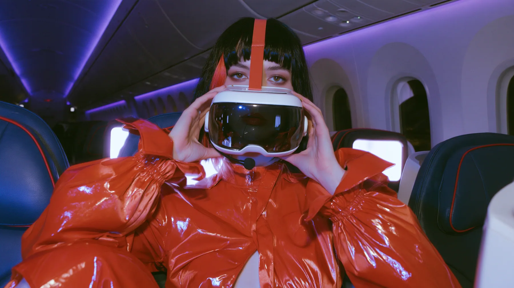 Jet-set fashion editorial, night flight: a medium close-up of a Caucasian American woman in her late twenties with pale skin and a glossy black blunt bob with heavy straight bangs and one clay-orange streak, a thin…