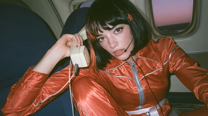 Jet-set fashion editorial, dusk flight: an eye-level medium shot of a Caucasian American woman in her early thirties, a grown woman with elegant mature features, an angular face and defined cheekbones, pale freckled…