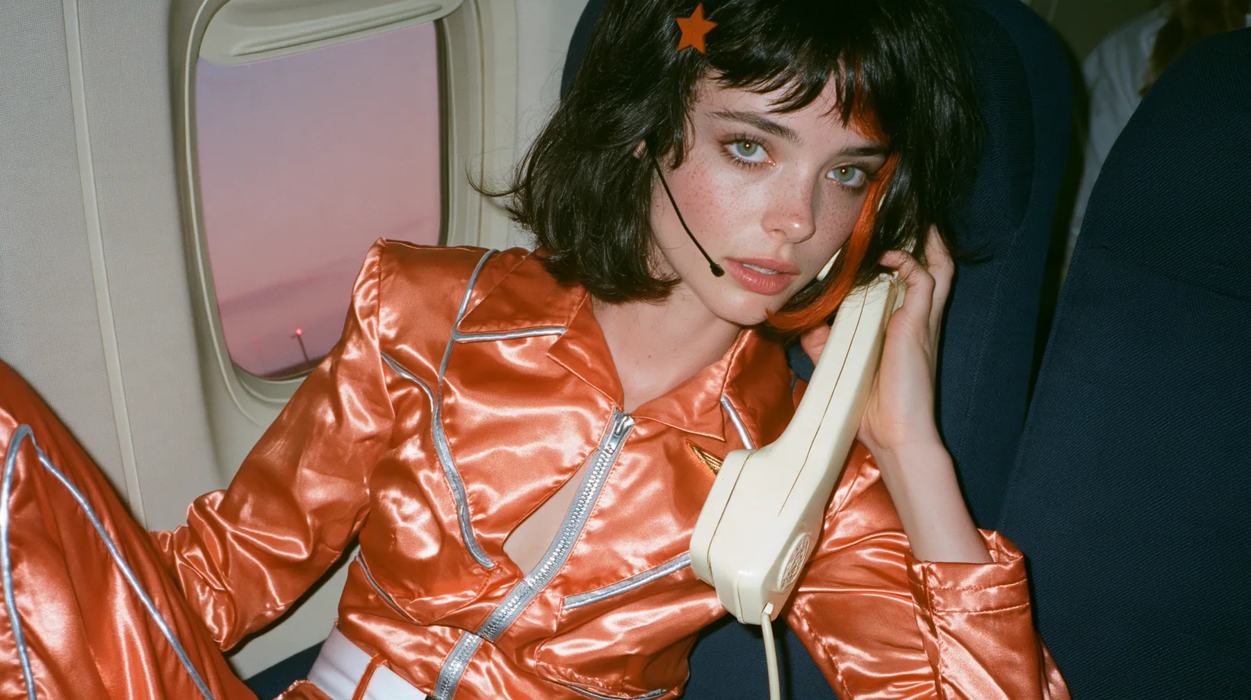Jet-set fashion editorial, dusk flight: an eye-level medium shot of a Caucasian American woman in her early thirties, a grown woman with elegant mature features, an angular face and defined cheekbones, pale freckled…