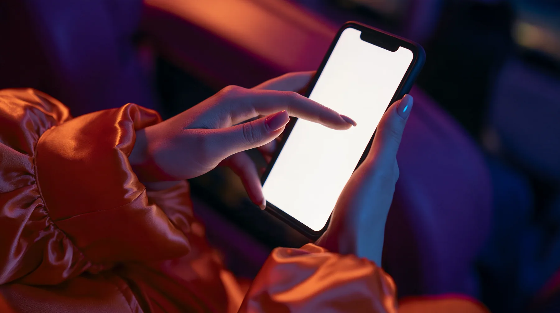 Editorial macro, night flight: a pale woman's left hand with short natural nails holds a black smartphone upright, its blank bright white screen toward the camera, the index finger of her right hand touching the…