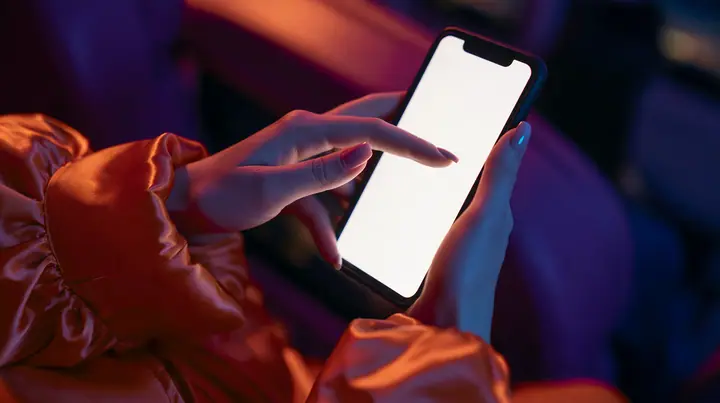 Editorial macro, night flight: a pale woman's left hand with short natural nails holds a black smartphone upright, its blank bright white screen toward the camera, the index finger of her right hand touching the…