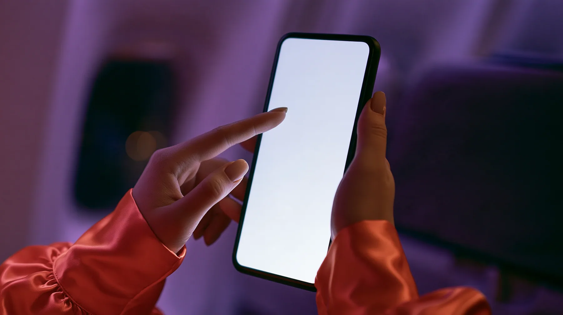 Editorial macro, night flight: a pale woman's left hand with short natural nails holds a black smartphone upright, its blank bright white screen toward the camera, the index finger of her right hand touching the…