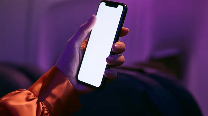 Editorial macro, night flight: a pale woman's hand with short natural unpainted nails, a glossy international-orange satin cuff at the wrist, holding a black smartphone upright, her thumb pressing the upper left of…