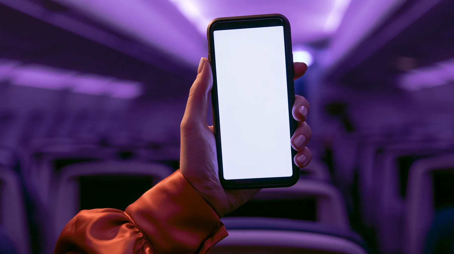 Editorial macro, night flight: a pale woman's hand with short natural unpainted nails, a glossy international-orange satin cuff at the wrist, holding a black smartphone upright, her thumb pressing the upper left of…