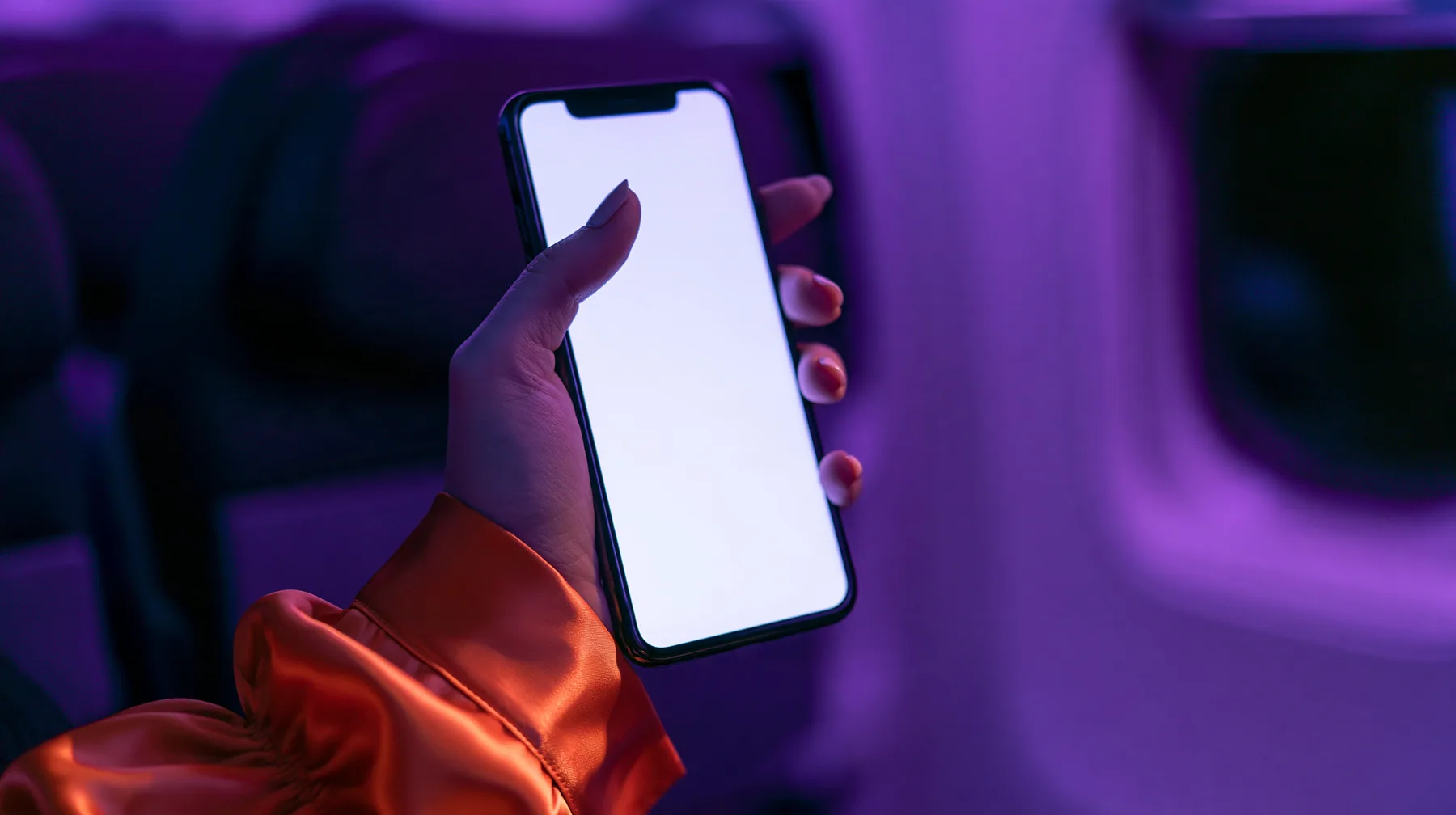 Editorial macro, night flight: a pale woman's hand with short natural unpainted nails, a glossy international-orange satin cuff at the wrist, holding a black smartphone upright, her thumb pressing the upper left of…