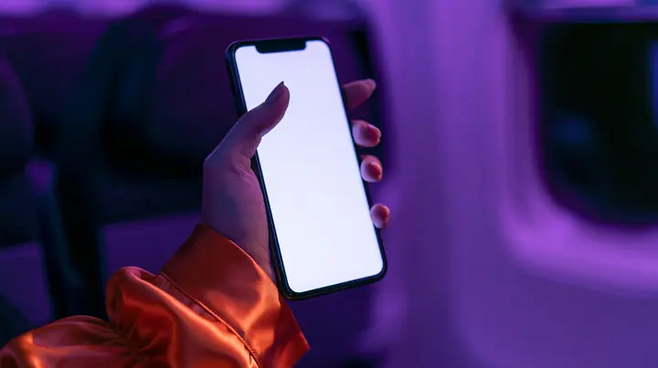Editorial macro, night flight: a pale woman's hand with short natural unpainted nails, a glossy international-orange satin cuff at the wrist, holding a black smartphone upright, her thumb pressing the upper left of…