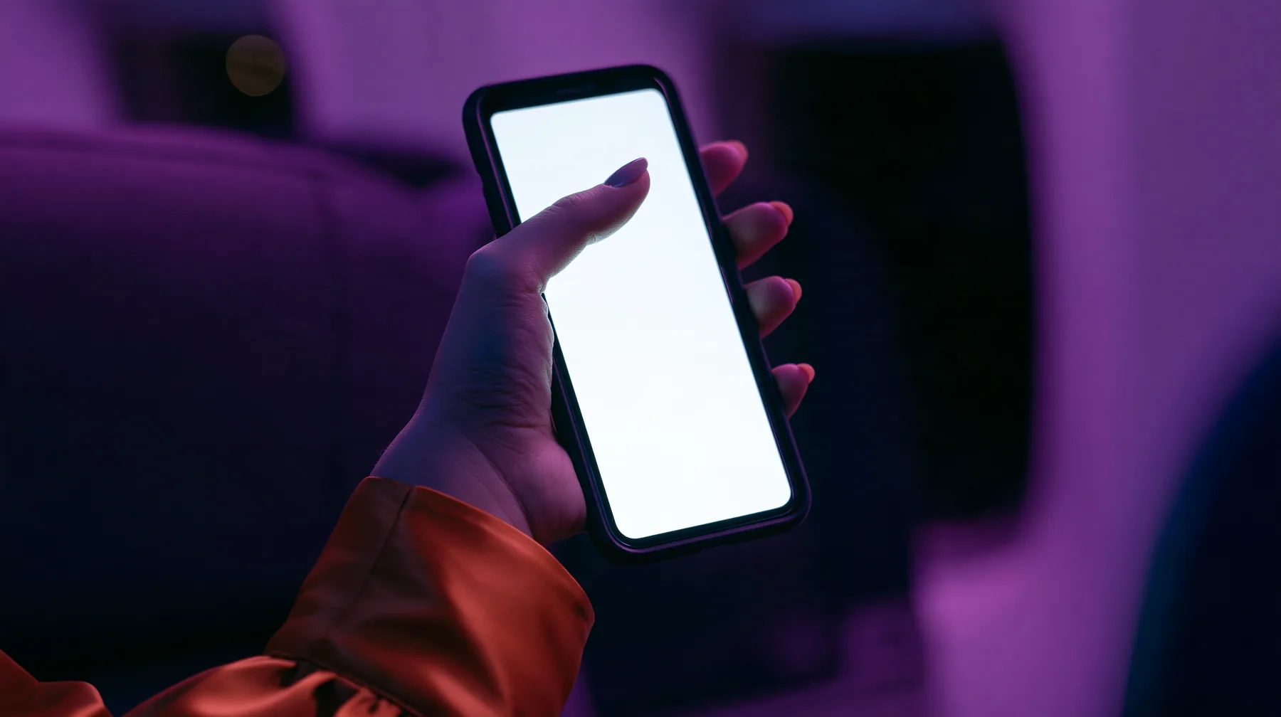Editorial macro, night flight: a pale woman's hand with short natural unpainted nails, a glossy international-orange satin cuff at the wrist, holding a black smartphone upright, her thumb pressing the upper left of…