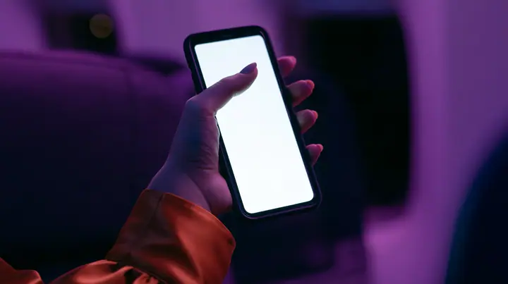 Editorial macro, night flight: a pale woman's hand with short natural unpainted nails, a glossy international-orange satin cuff at the wrist, holding a black smartphone upright, her thumb pressing the upper left of…