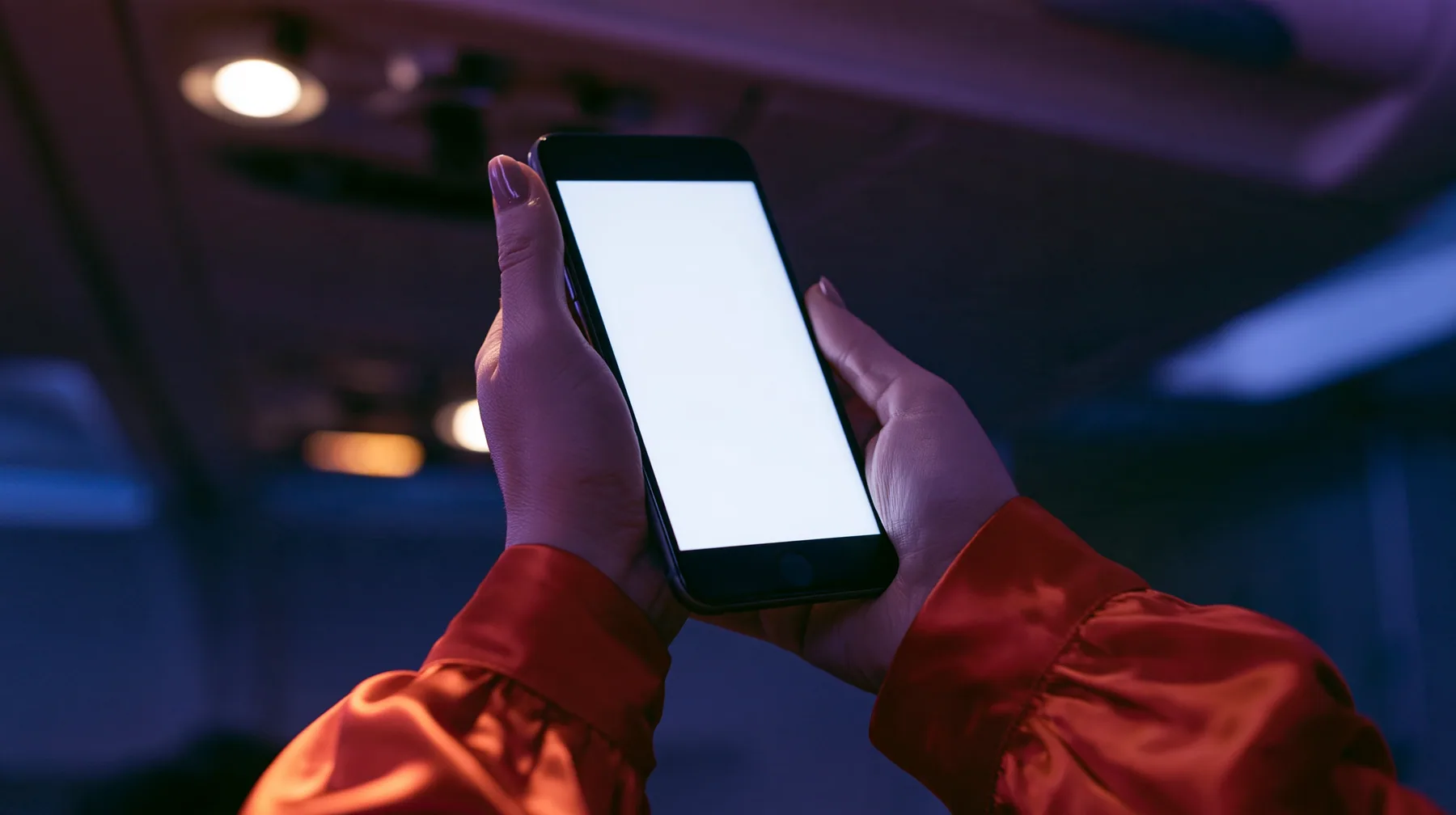 Editorial close-up, night flight: a fair-skinned hand, short clean nails, glossy international-orange satin sleeve cuff, holding a black smartphone in portrait at chest height, the thumb resting on the top left…