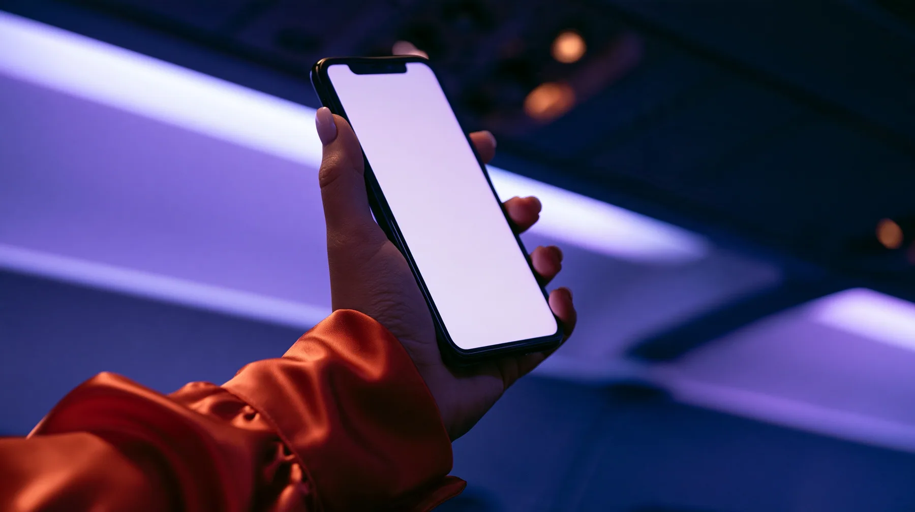Editorial close-up, night flight: a fair-skinned hand, short clean nails, glossy international-orange satin sleeve cuff, holding a black smartphone in portrait at chest height, the thumb resting on the top left…