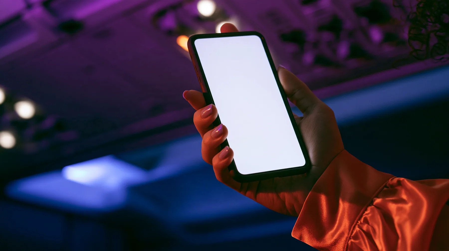 Editorial close-up, night flight: a fair-skinned hand, short clean nails, glossy international-orange satin sleeve cuff, holding a black smartphone in portrait at chest height, the thumb resting on the top left…