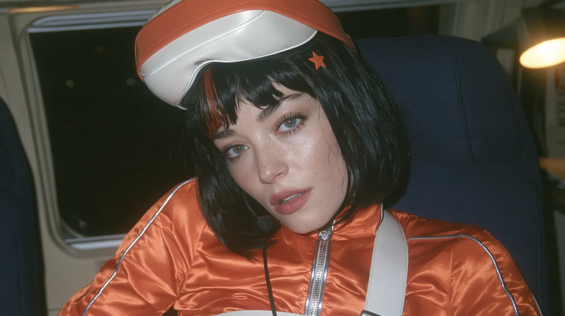Jet-set fashion editorial, night flight: an eye-level close-up of a Caucasian American woman in her early thirties, a grown woman with elegant mature features, an angular face and defined cheekbones, pale freckled…