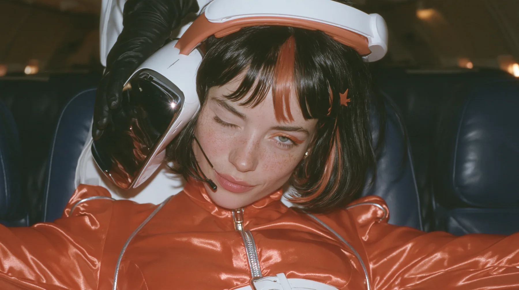 Close-up film still: a Caucasian woman in her early thirties with an angular adult face, pale freckled skin, a glossy black blunt bob with heavy bangs and one clay-orange streak, a small orange star hair clip, a thin…