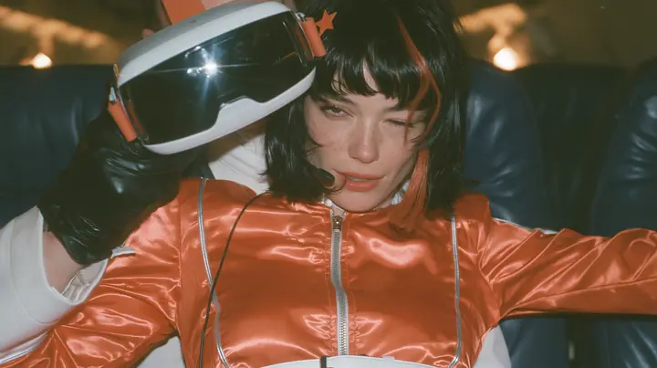 Close-up film still: a Caucasian woman in her early thirties with an angular adult face, pale freckled skin, a glossy black blunt bob with heavy bangs and one clay-orange streak, a small orange star hair clip, a thin…