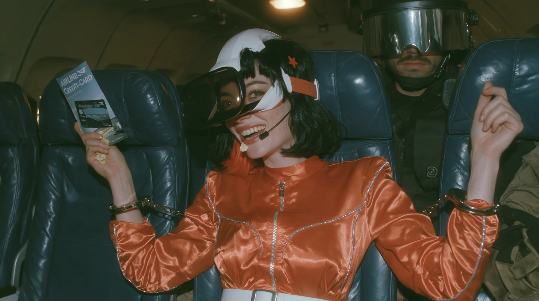 Cinematic film still, night airliner cabin with navy leather seats, all of them empty: a Caucasian woman in her early thirties with an angular adult face, pale freckled skin, a glossy black blunt bob with heavy bangs…