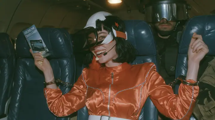 Cinematic film still, night airliner cabin with navy leather seats, all of them empty: a Caucasian woman in her early thirties with an angular adult face, pale freckled skin, a glossy black blunt bob with heavy bangs…
