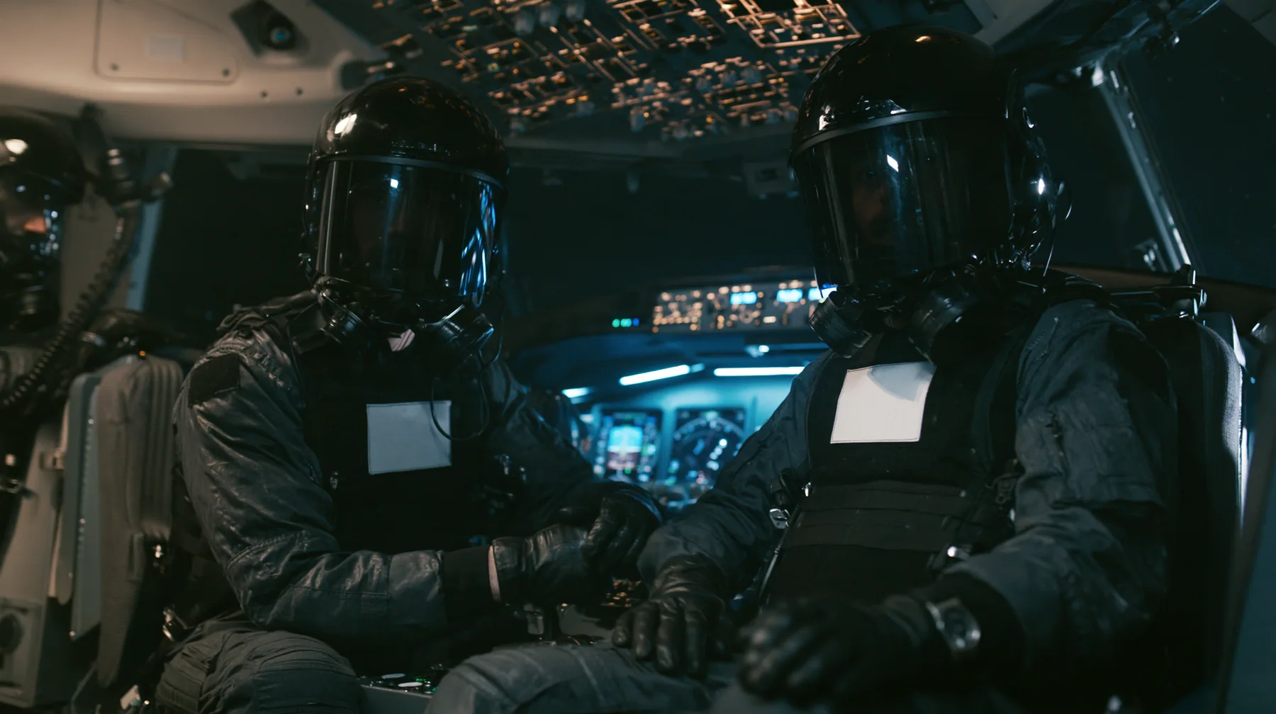 Cinematic film still inside a modern airliner cockpit at night: two guards in matte-black tactical gear and black helmets with mirrored visors hiding their faces, plain white chest patches, black gloves, sit in the…