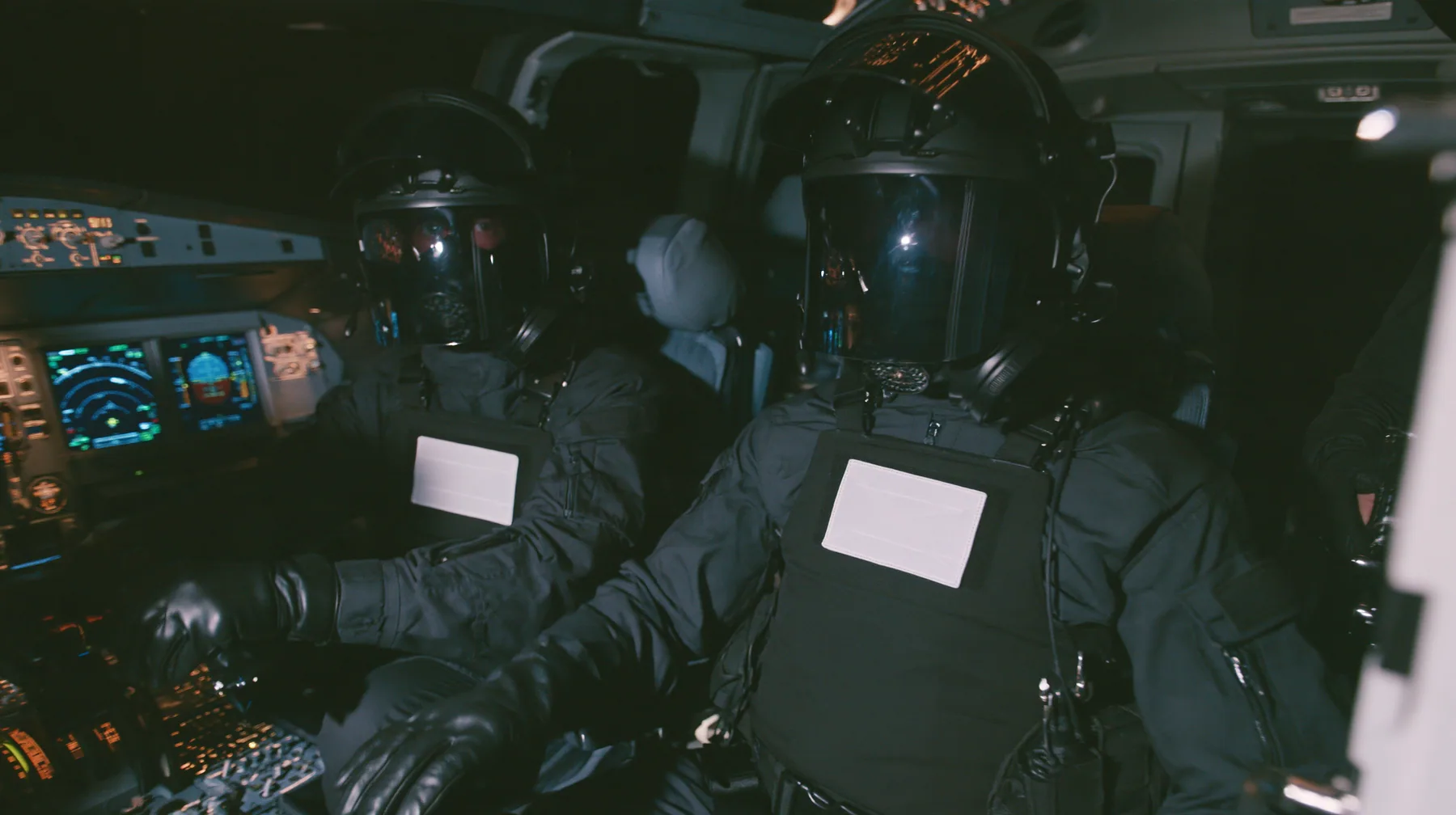 Cinematic film still inside a modern airliner cockpit at night: two guards in matte-black tactical gear and black helmets with mirrored visors hiding their faces, plain white chest patches, black gloves, sit in the…