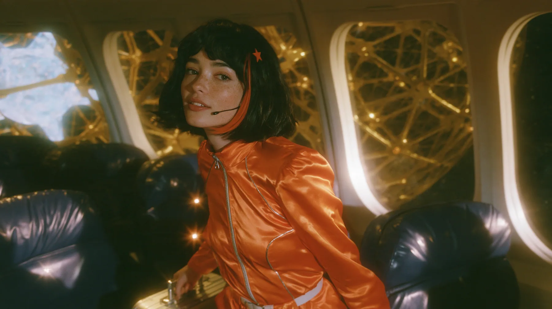 Cinematic film still: a Caucasian woman in her early thirties with an angular adult face, pale freckled skin, a glossy black blunt bob with heavy bangs and one clay-orange streak, a small orange star hair clip, a…
