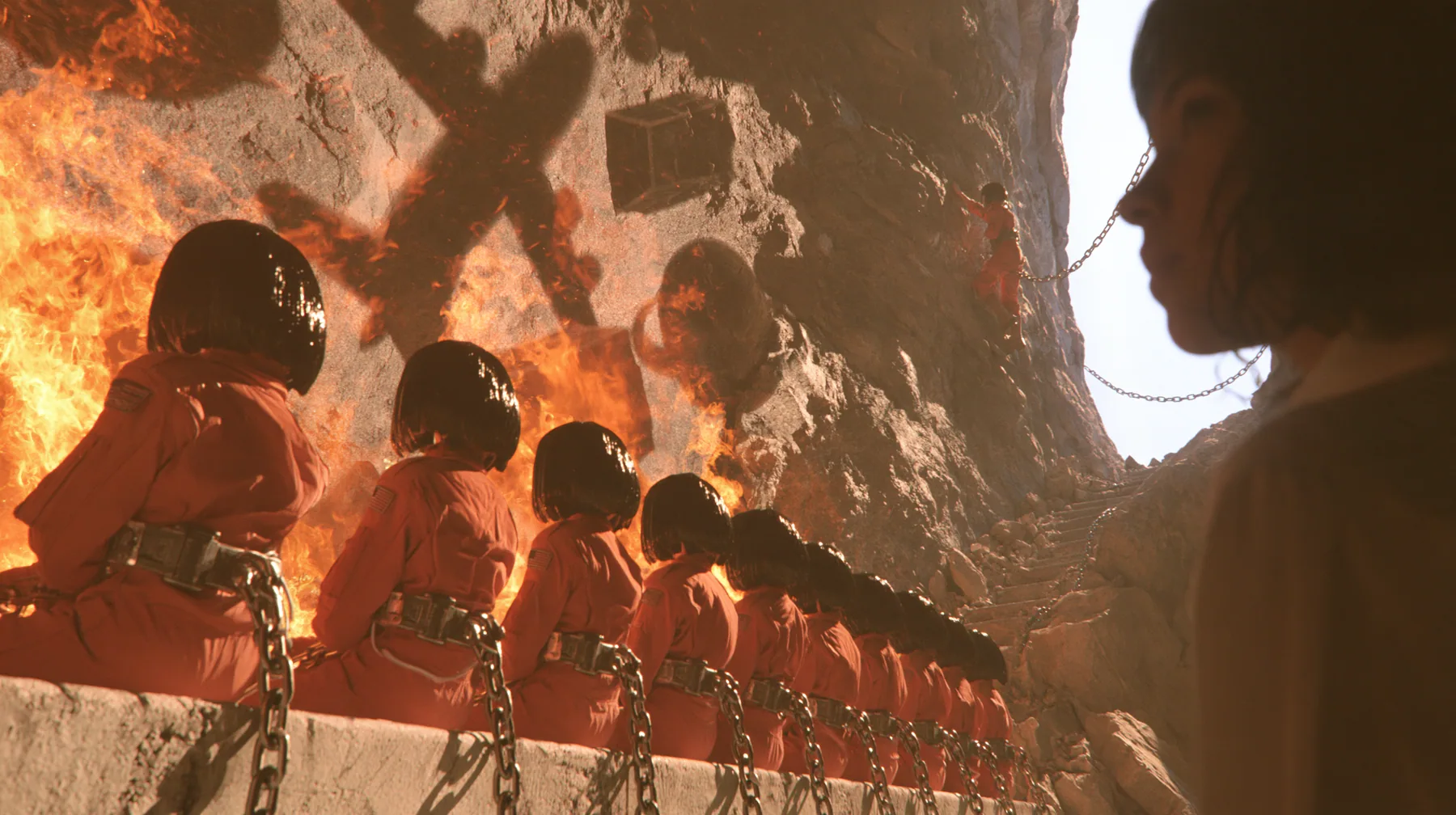Cinematic wide shot inside Plato's cave: a row of young women with glossy black bobs in orange flight suits sit chained on a stone bench facing a rock wall, watching huge dancing shadows of airplanes, boxes and…