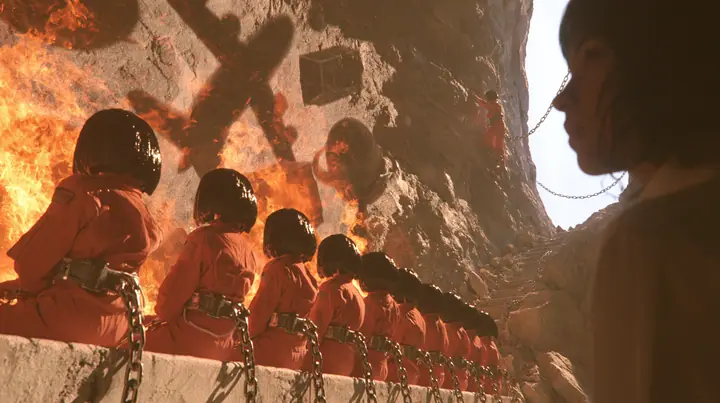 Cinematic wide shot inside Plato's cave: a row of young women with glossy black bobs in orange flight suits sit chained on a stone bench facing a rock wall, watching huge dancing shadows of airplanes, boxes and…