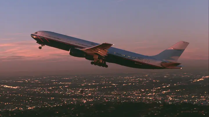 Cinematic exterior shot: a white airliner with a navy belly and a thin red and navy cheat-line climbing steeply out of a city at dusk, city lights below, pink and orange dusk sky, the camera slightly below and beside…