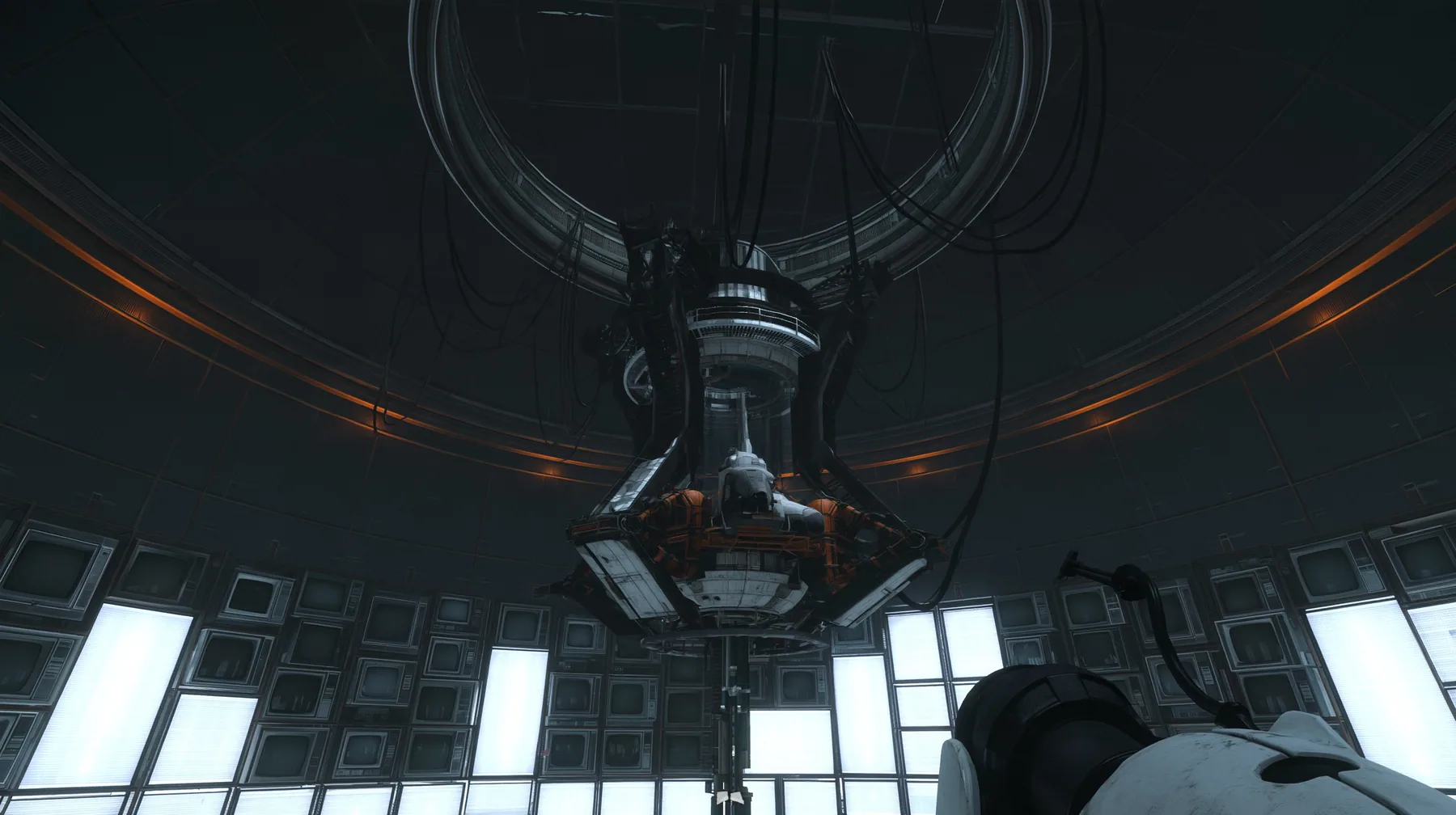 the same dark circular AI chamber with the ceiling ring and hanging cables, its curved walls now lined floor to ceiling with dozens of old CRT monitors and flat panel screens glowing faint white, warm amber accent…