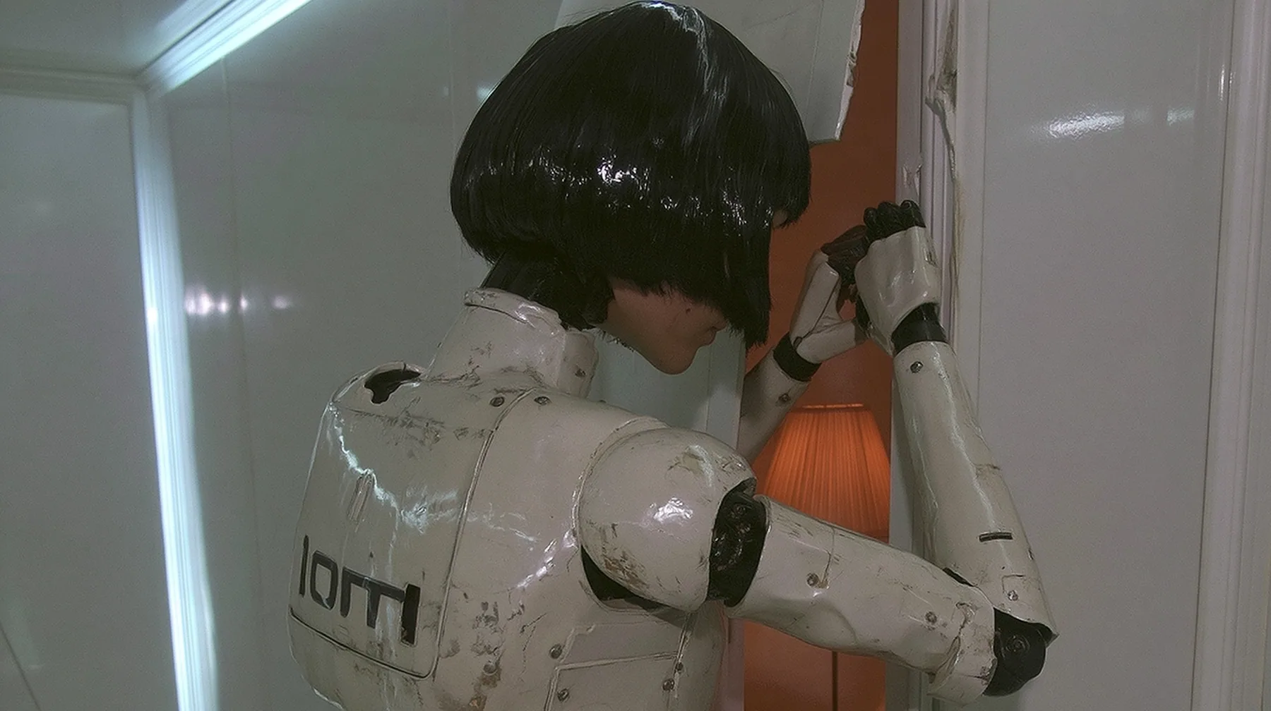 over-the-shoulder from behind at shoulder height: a scuffed slender porcelain humanoid android of glossy white plating with black joints, the back of her glossy black blunt bob toward the camera, grips the edge of a…