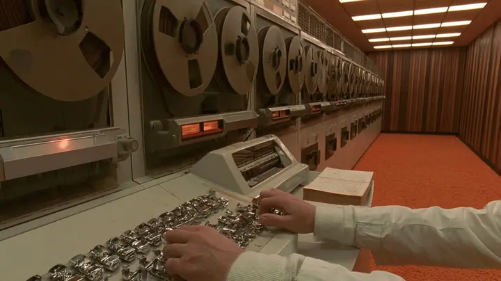 cinematic console-height shot along a row of 1960s reel-to-reel tape drives receding into a wood-panelled computer room with orange carpet: in the foreground a scientist's hands in white lab-coat cuffs on a row of…