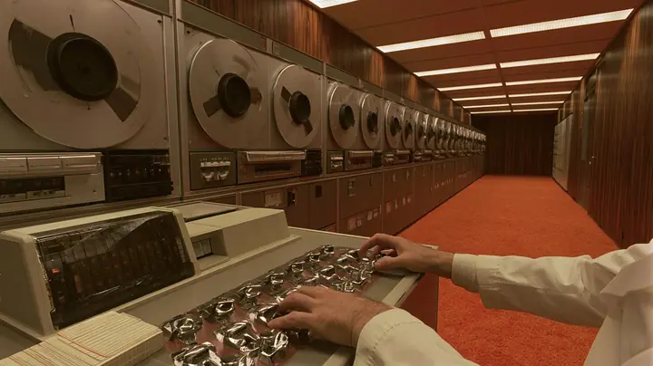 cinematic console-height shot along a row of 1960s reel-to-reel tape drives receding into a wood-panelled computer room with orange carpet: in the foreground a scientist's hands in white lab-coat cuffs on a row of…