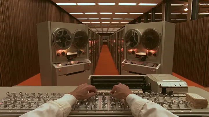 cinematic console-height shot along a row of 1960s reel-to-reel tape drives receding into a wood-panelled computer room with orange carpet: in the foreground a scientist's hands in white lab-coat cuffs on a row of…