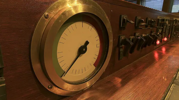 cinematic close-up of a big brass gauge on a polished wooden 1960s console: a cream dial with a green zone at the left and a red zone at the upper right, plain tick marks, the black needle trembling deep in the red,…
