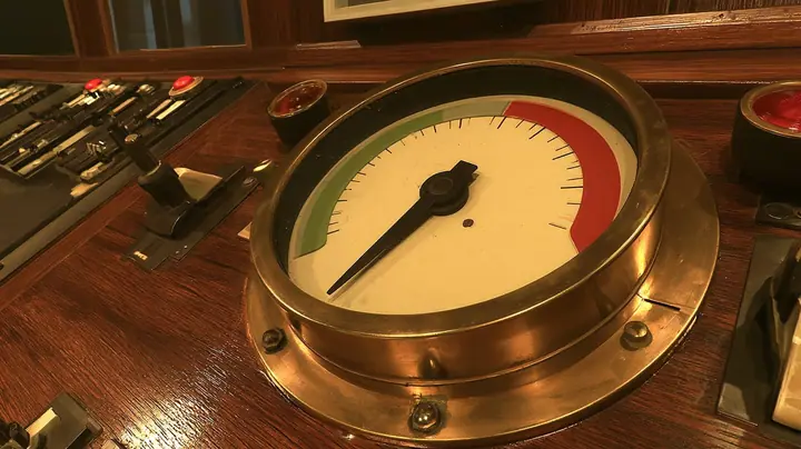 cinematic close-up of a big brass gauge on a polished wooden 1960s console: a cream dial with a green zone at the left and a red zone at the upper right, plain tick marks, the black needle trembling deep in the red,…