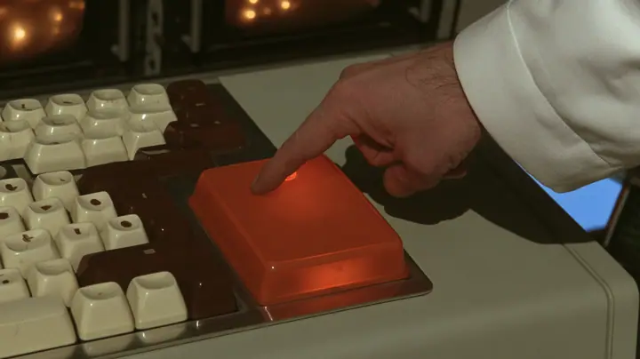 Macro cinematic film still at keyboard height on a chunky 1970s Aperture control console: rows of cream and brown keys with rounded tops and, in the middle, one oversized square key of glossy orange plastic glowing…
