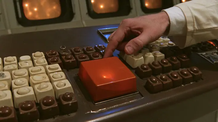Macro cinematic film still at keyboard height on a chunky 1970s Aperture control console: rows of cream and brown keys with rounded tops and, in the middle, one oversized square key of glossy orange plastic glowing…