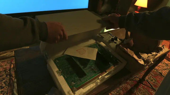 Top-down cinematic film still onto a low wooden coffee table at night: two hands in dark grey hoodie sleeves lift the lid off a sleek brand-new plain white game console nested in packing foam, revealing a green…