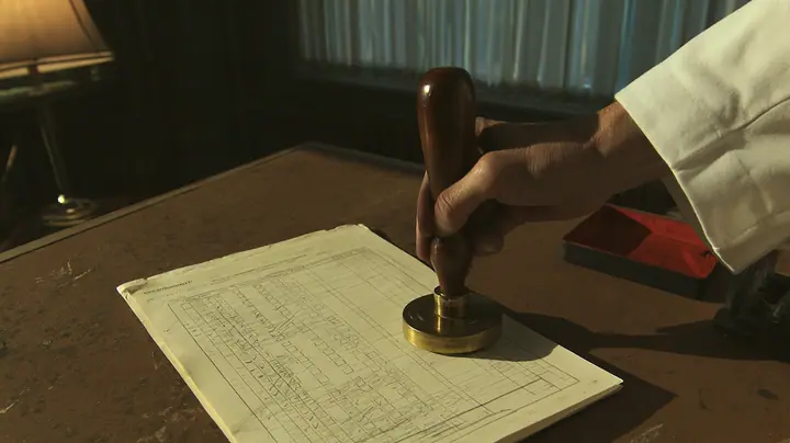 Macro cinematic film still at desk level in a 1970s Aperture office: an official cream evaluation form with rows of pencil-ticked boxes lies on a worn brown desk, and above it, filling the top of the frame, a big…