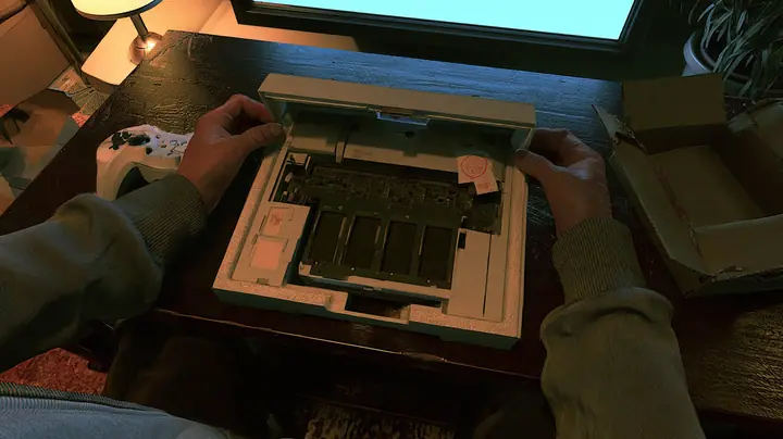 Top-down cinematic film still: on a dark wooden coffee table hands in a grey hoodie pull the cover off a new white game console in its foam tray, its circuit board bare with empty memory slots and a little paper slip…