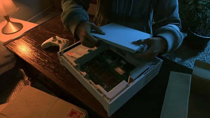 Top-down cinematic film still: on a dark wooden coffee table hands in a grey hoodie pull the cover off a new white game console in its foam tray, its circuit board bare with empty memory slots and a little paper slip…