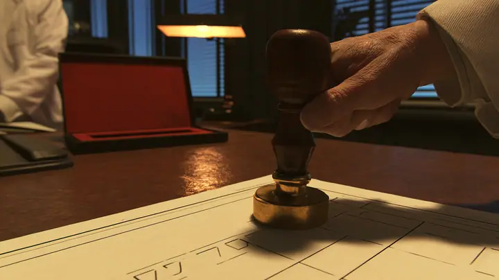 Macro cinematic film still: a heavy wood-handled rubber stamp with a brass collar, held by a hand in a white lab coat sleeve, hovers over a cream form with empty tick boxes on a polished brown desk, an open red ink…