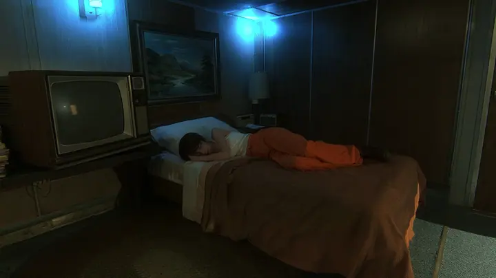 cinematic still of a small windowless 1970s motel-like room inside a test facility at night: a woman with dark brown hair, a white tank top and an orange jumpsuit sleeps on her side facing the camera on a single bed…