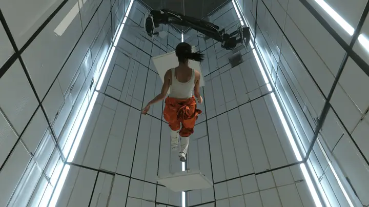 cinematic film still from the floor looking up a towering white test chamber of black-seamed panels and vertical light strips: high above, a woman with a dark ponytail, white tank top and orange jumpsuit knotted at…