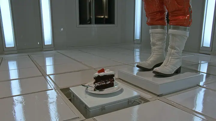 macro film still at floor level in a bright white test chamber: a single slice of black forest cake on a white plate with a small fork rides up on a little white floor panel lifting out of the tiles, a woman's orange…