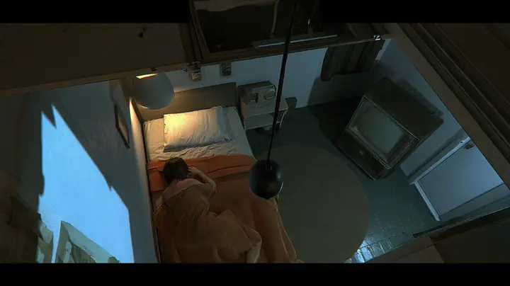 overhead film still looking straight down into a small motel-like room at night: a bed with a brown blanket and white pillow, a woman curled asleep with only her dark hair visible, beside her the top of a…