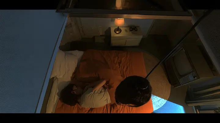 overhead film still looking straight down into a small motel-like room at night: a bed with a brown blanket and white pillow, a woman curled asleep with only her dark hair visible, beside her the top of a…