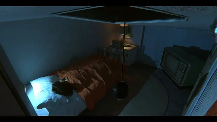 overhead film still looking straight down into a small motel-like room at night: a bed with a brown blanket and white pillow, a woman curled asleep with only her dark hair visible, beside her the top of a…