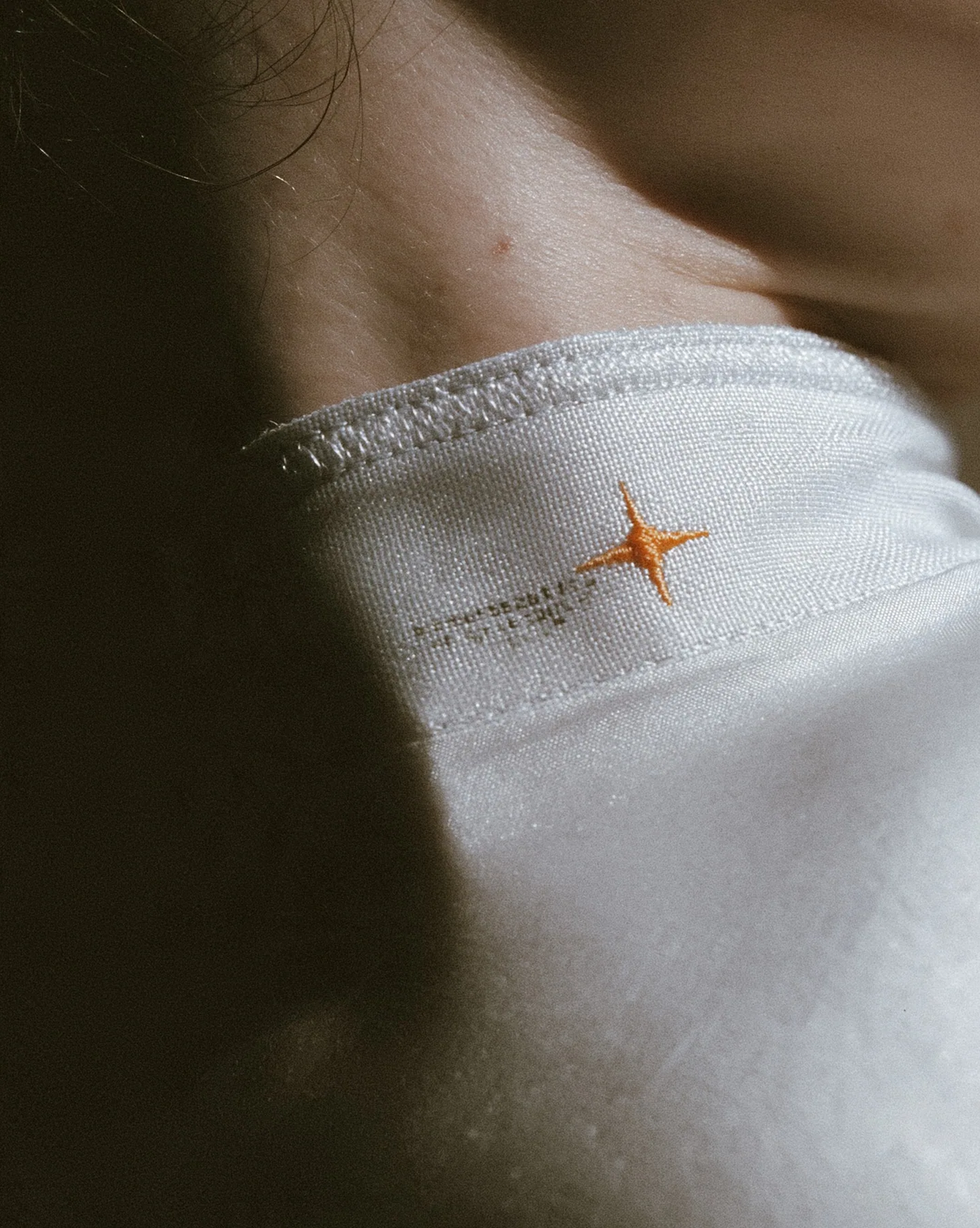 macro fashion-editorial close-up of a young woman's collarbone and jaw, a stitched technical woven label with tiny serial codes and a clay-orange eight-pointed spark on matte white ripstop, razor-thin focus, soft…