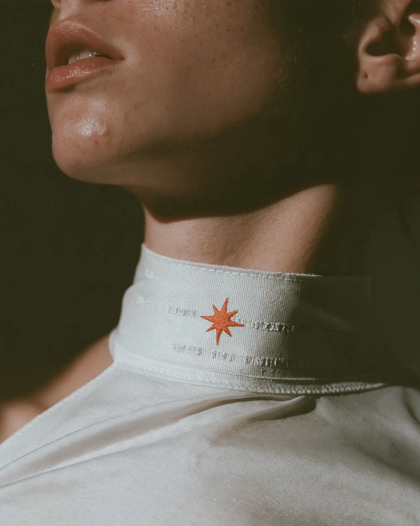 macro fashion-editorial close-up of a young woman's collarbone and jaw, a stitched technical woven label with tiny serial codes and a clay-orange eight-pointed spark on matte white ripstop, razor-thin focus, soft…