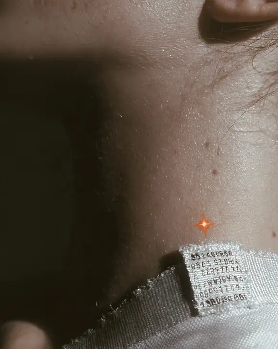 macro fashion-editorial close-up of a young woman's collarbone and jaw, a stitched technical woven label with tiny serial codes and a clay-orange eight-pointed spark on matte white ripstop, razor-thin focus, soft…